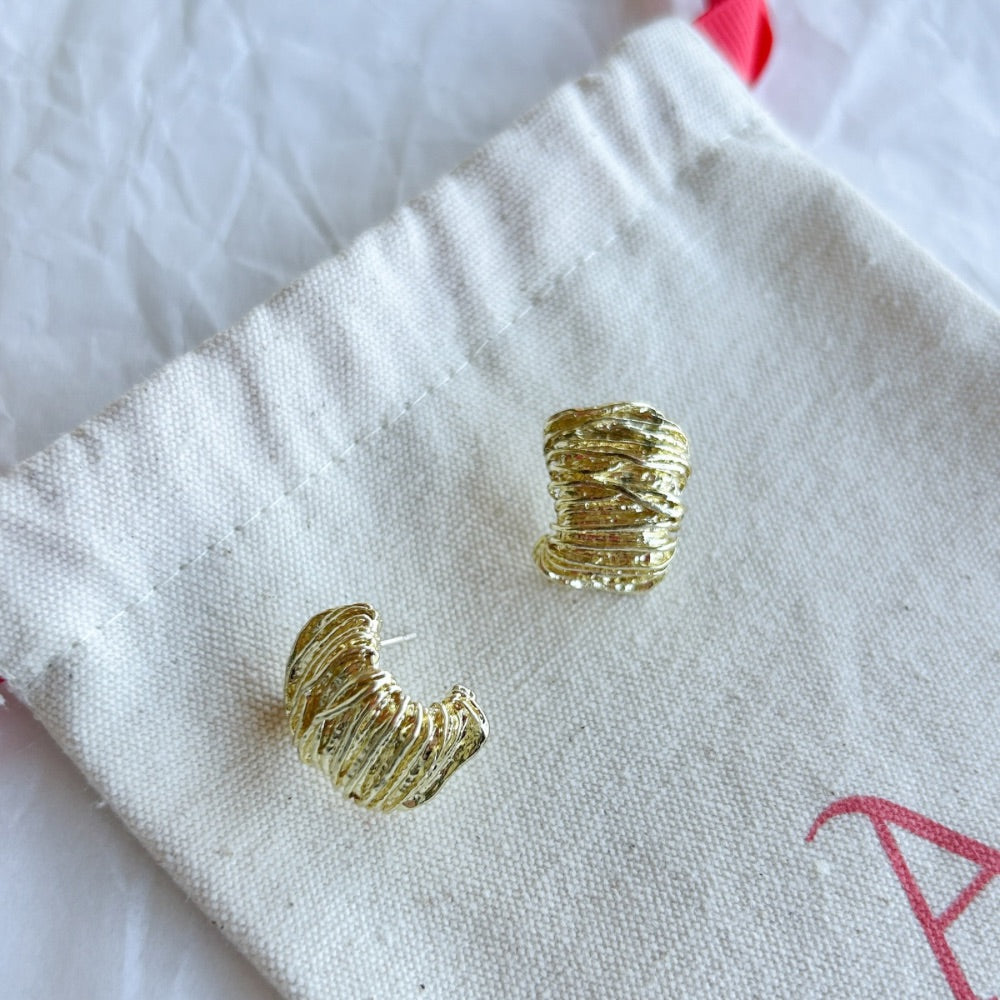 Aros Dorados Bergen con textura entrelazada y acabado en oro mate, exhibidos sobre una bolsa de tela Altapinta.