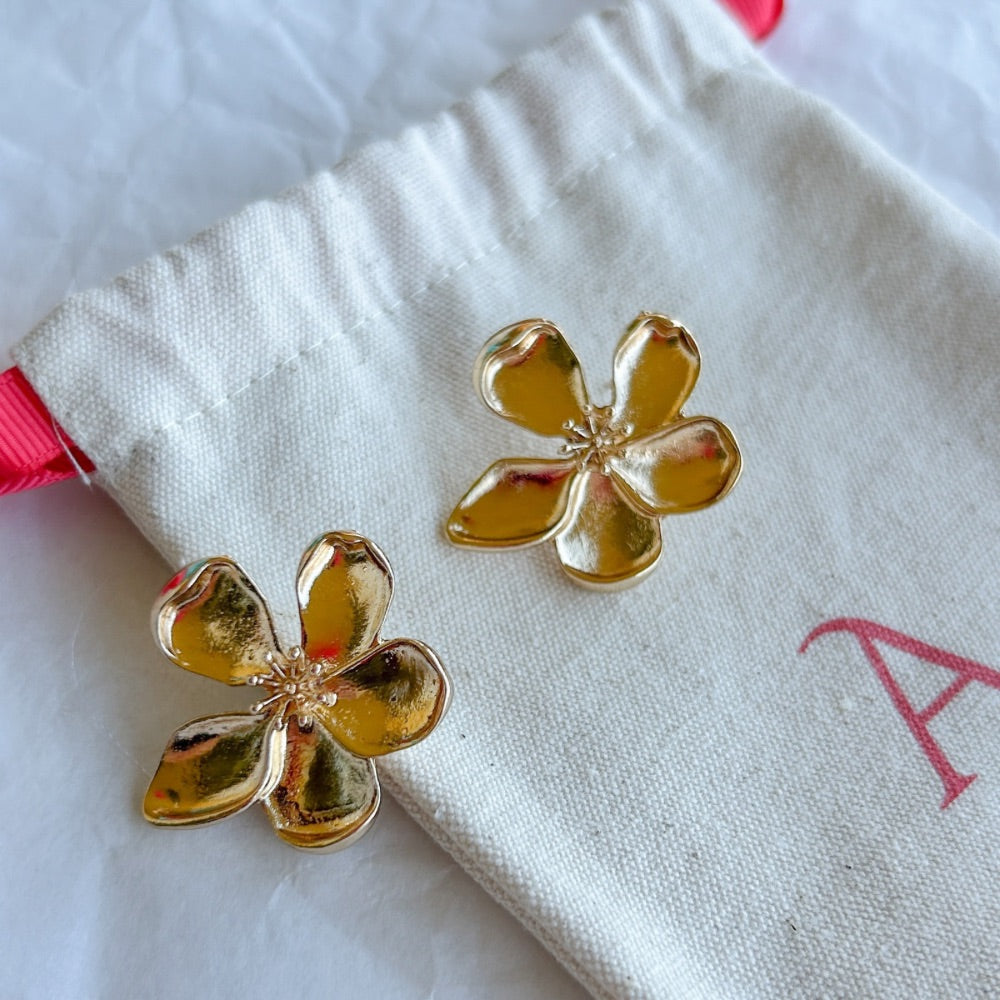 Aros Dorados Dublín con diseño floral de cinco pétalos y textura sutil, exhibidos sobre una bolsa de tela clara con detalles en rojo.
