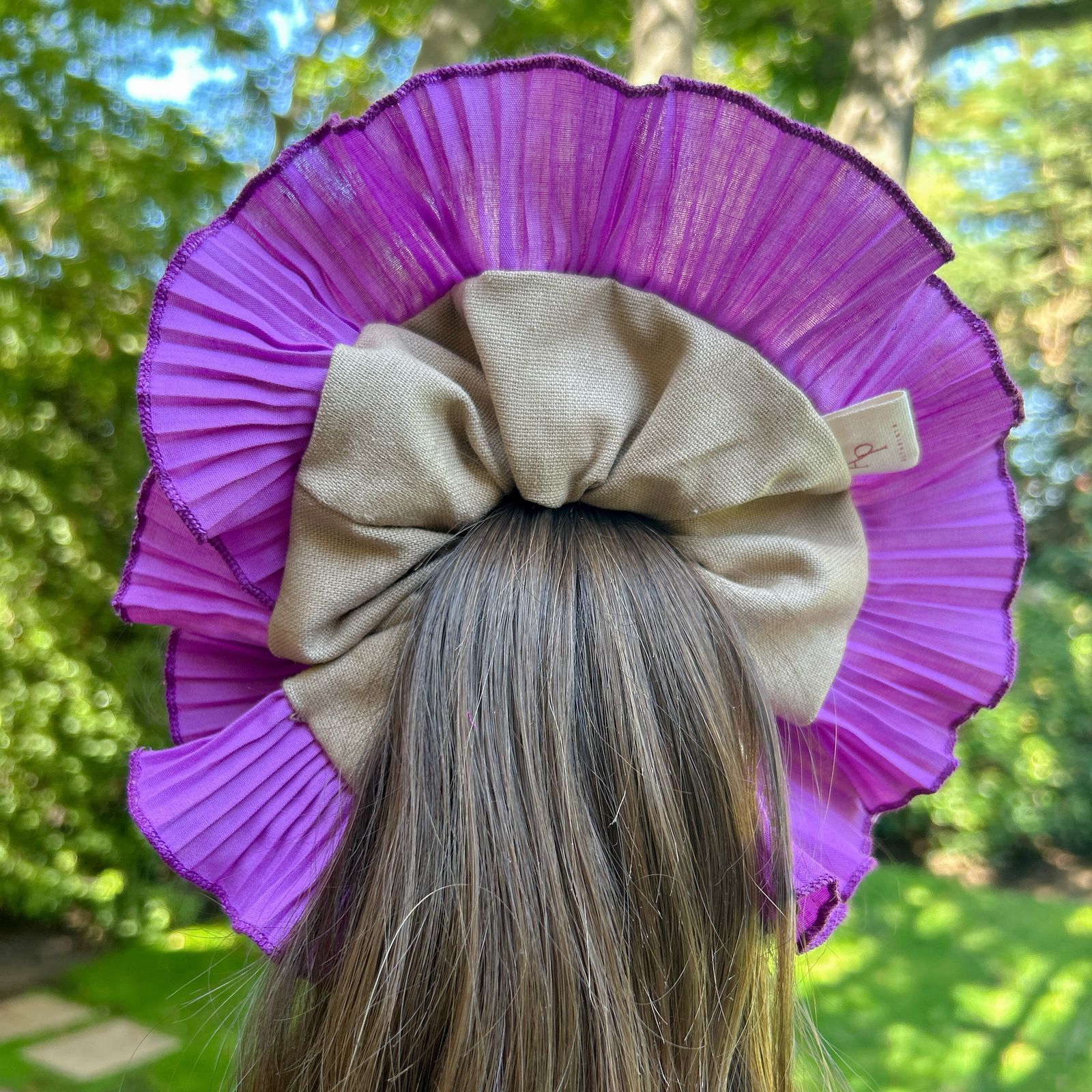 Bamba Simple Arena con lazo beige y plisado morado, adornando una coleta alta en un entorno natural.