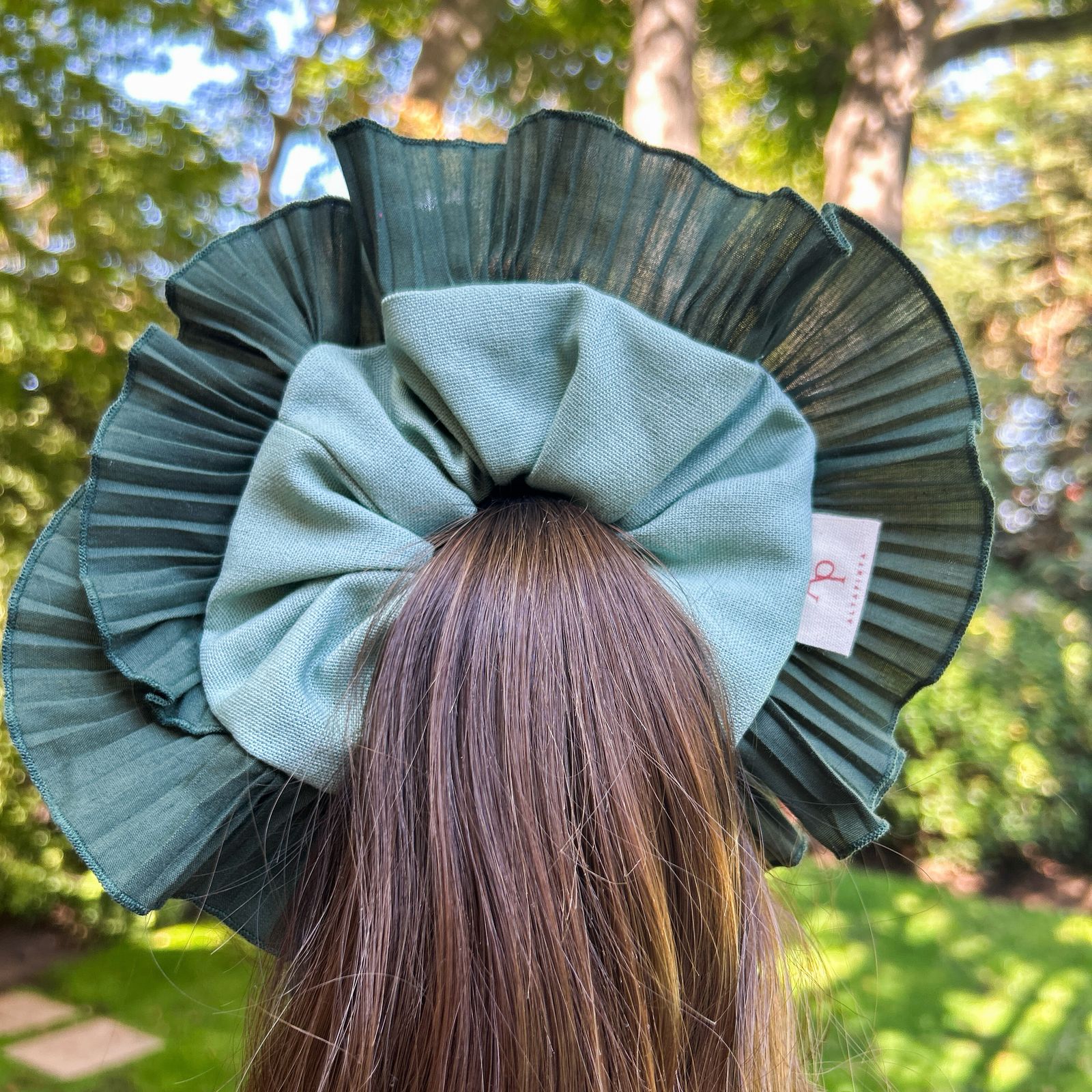 Bamba Simple Selva con lazo verde y plisado estructurado en verde oscuro, adornando una coleta alta en un entorno natural.