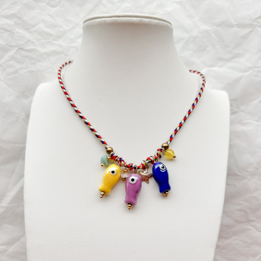 Collar Artesanal Olas con tejido trenzado en rojo, blanco y azul, decorado con dijes de peces de cerámica y detalles dorados, exhibido sobre un busto blanco.