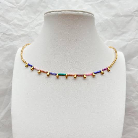 Collar Artesanal Raíz con base dorada, mostacillas en tonos vibrantes y detalles dorados en forma de gotas, exhibido sobre un busto blanco.