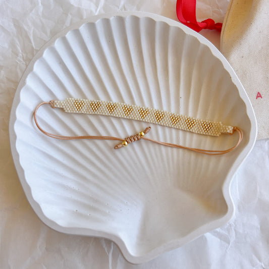 Pulsera Artesanal Corazones Amelia en tonos dorados, con diseño de corazones tejidos en mostacillas sobre una base blanca.