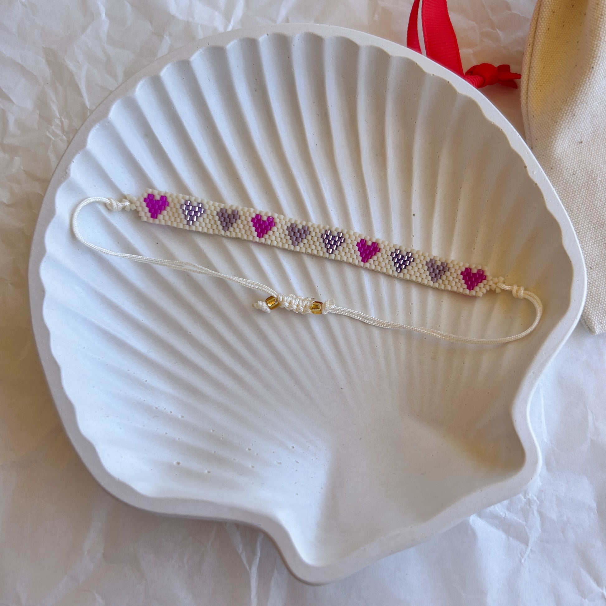 Pulsera Artesanal Corazones Amelia en tonos lilas y rosas, tejida a mano con mostacillas y cierre ajustable sobre una concha blanca.