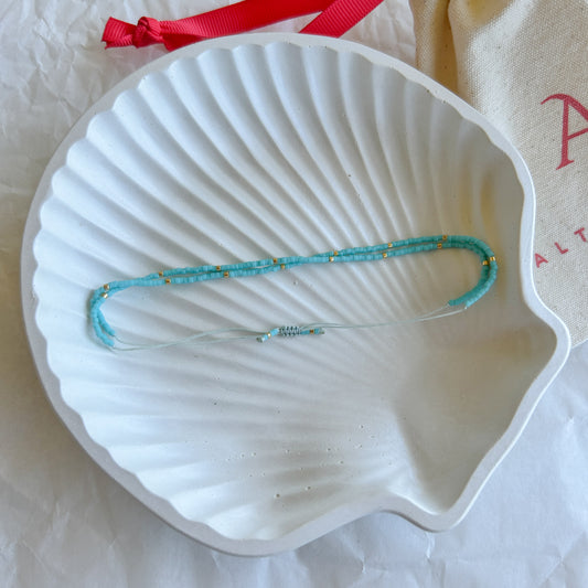 Pulsera artesanal doble con cuentas en tonos turquesa y detalles dorados, presentada sobre un plato en forma de concha.