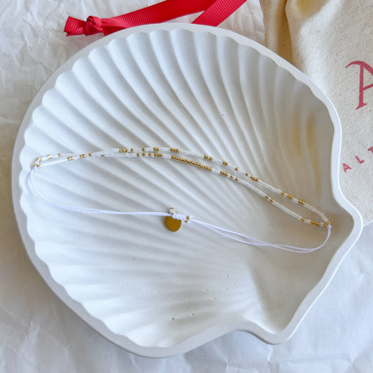 Pulsera artesanal doble en tonos blanco y dorado, exhibida sobre un plato en forma de concha.