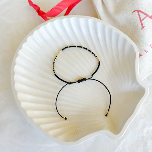 Pulsera artesanal simple Augusta con cuentas en negro y dorado, cierre ajustable en hilo negro, exhibida en un plato de concha blanca.