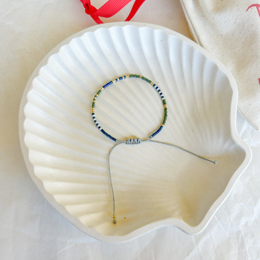 Pulsera Artesanal Simple Lourdes sobre una concha blanca, con mostacillas en tonos azul, verde y dorado, y un hilo gris ajustable. Accesorio boho chic elegante y minimalista.