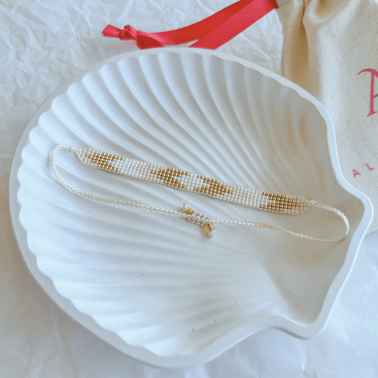 Pulsera Artesanal Tejida de Mostacillas Agustina en tonos blanco y dorado sobre una concha blanca, destacando su diseño minimalista con cierre ajustable.