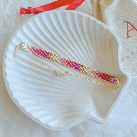 Pulsera Artesanal Tejida de Mostacillas Agustina en rosa y fucsia sobre una concha blanca, mostrando su diseño femenino con detalles en dorado.