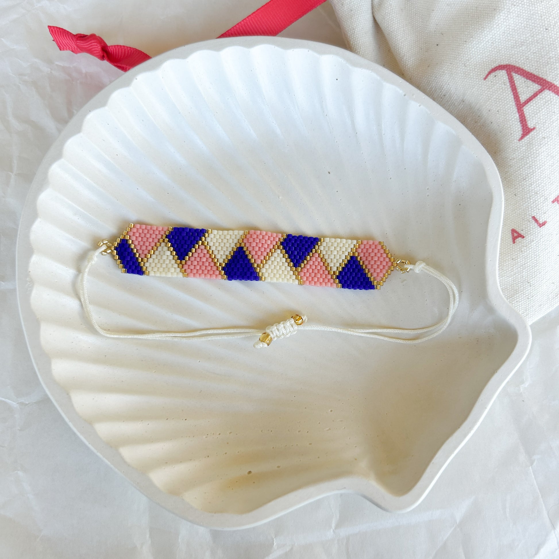 Pulsera Artesanal Tejida de Mostacillas Amanda sobre una concha blanca, mostrando su diseño de triángulos en azul, rosa y crema con detalles dorados y cierre ajustable.