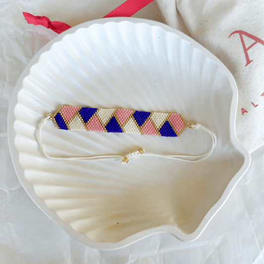 Pulsera Artesanal Tejida de Mostacillas Amanda sobre una concha blanca, mostrando su diseño de triángulos en azul, rosa y crema con detalles dorados y cierre ajustable.