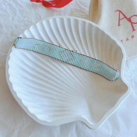 Pulsera Artesanal Tejida de Mostacillas Antonia en tonos pastel con detalles dorados, exhibida sobre una concha blanca.
