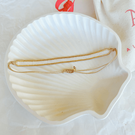 Pulsera Artesanal Tejida de Mostacillas Aurora en color blanco con dorado, colocada sobre una concha blanca, mostrando su estilo elegante y tejido artesanal de mostacillas con detalles dorados.