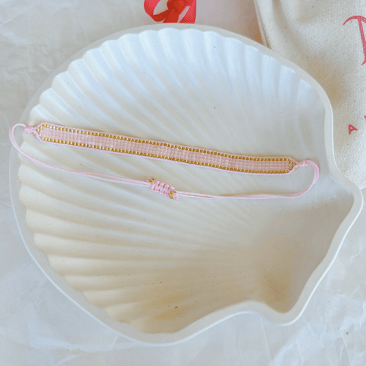 Pulsera Artesanal Tejida de Mostacillas Aurora en color rosa con dorado, exhibida sobre una concha blanca, resaltando su diseño delicado y sofisticado con cierre ajustable.