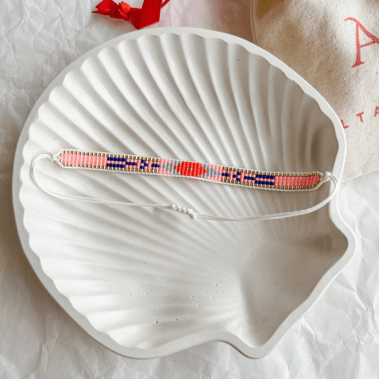Pulsera Artesanal Tejida de Mostacillas Belén en tonos coral, azul y dorado, exhibida sobre una concha blanca, destacando su diseño detallado y cierre ajustable.