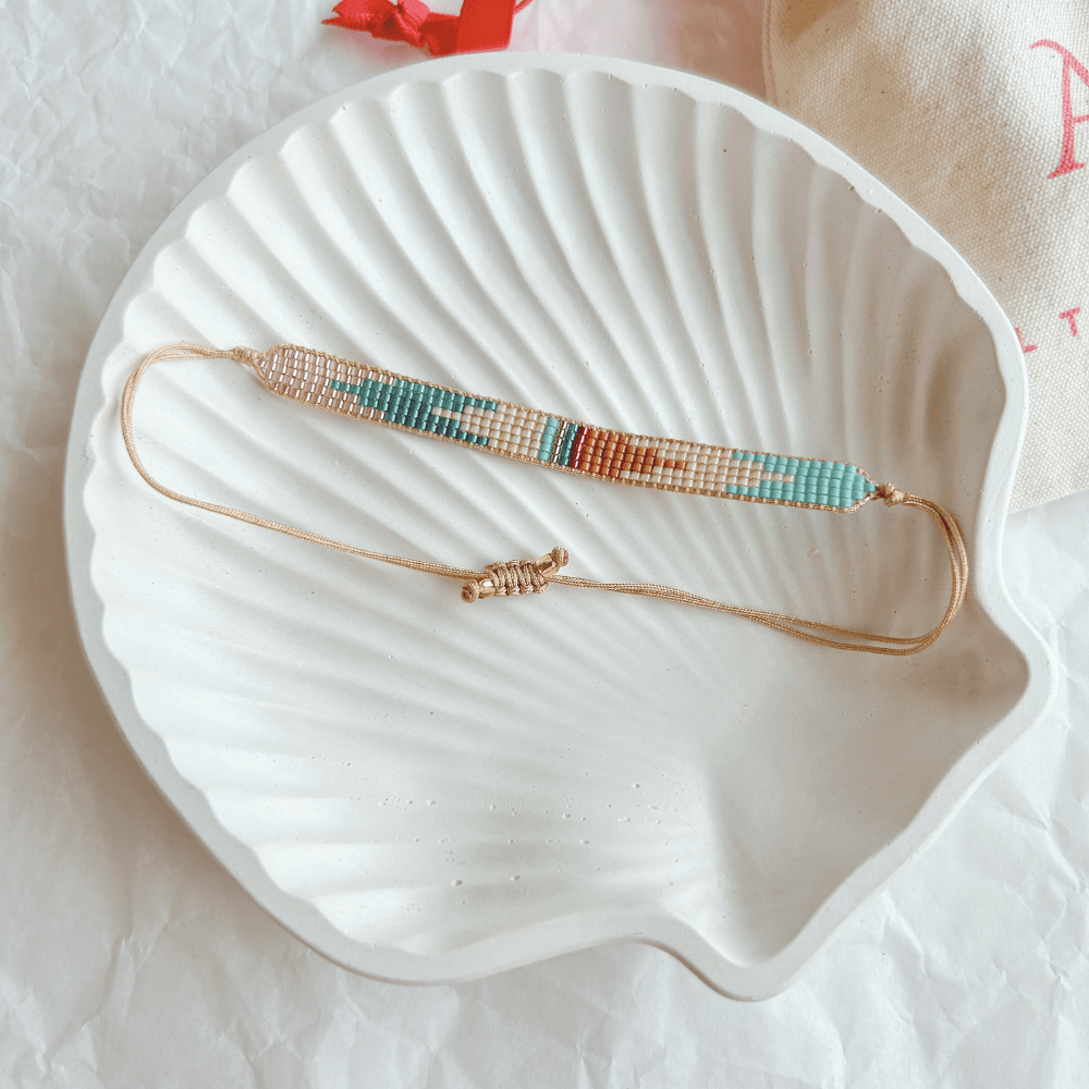 Pulsera Artesanal Tejida de Mostacillas Cristina en tonos multicolor con detalles dorados, ideal para un look boho-chic, sobre una concha blanca.