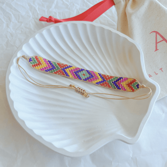 Pulsera Artesanal Tejida de Mostacillas Emilia con patrón geométrico colorido en tonos rojo, azul, verde, amarillo y morado, sobre una concha blanca, con cierre ajustable.