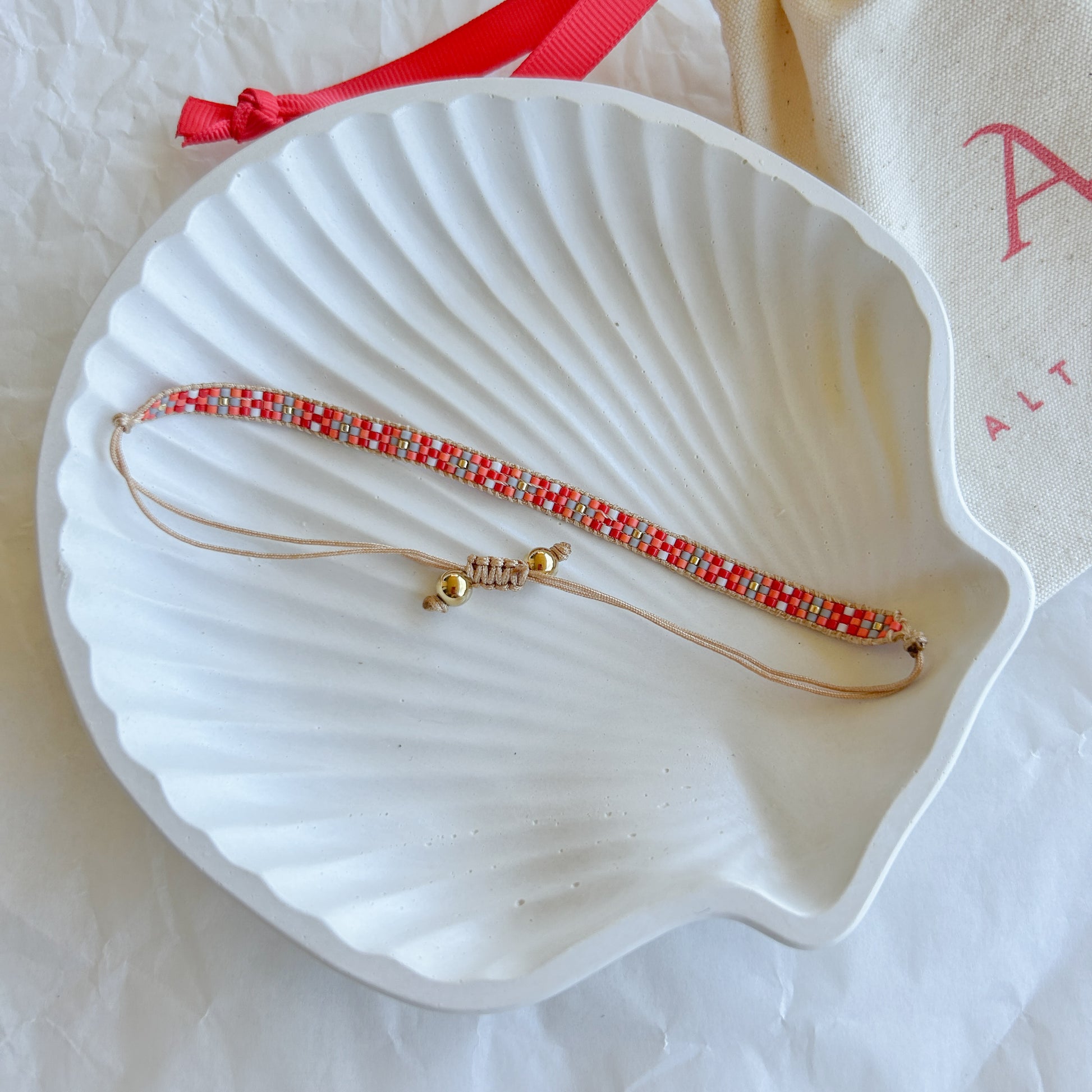 Pulsera Artesanal Tejida de Mostacillas Fernanda con patrón en tonos rojo, blanco y dorado, sobre una concha blanca, con cierre ajustable.