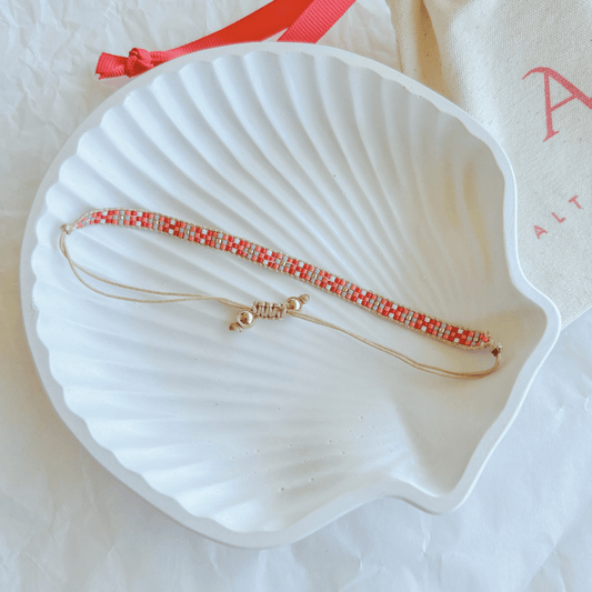 Pulsera Artesanal Tejida de Mostacillas Fernanda con patrón en tonos rojo, blanco y dorado, sobre una concha blanca, con cierre ajustable.