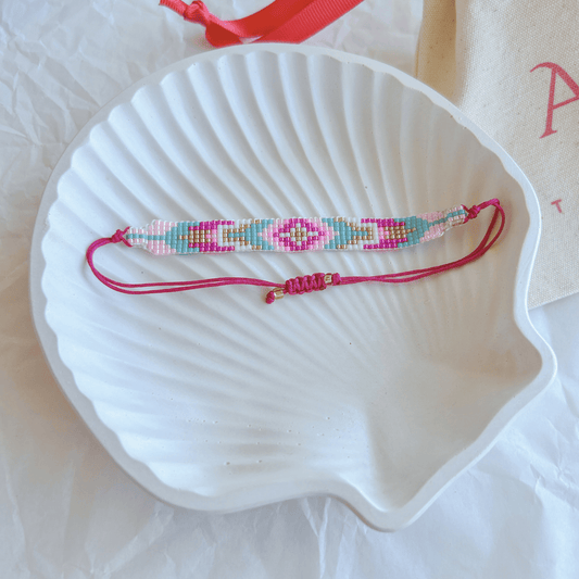 Pulsera Artesanal Tejida de Mostacillas Florencia con diseño geométrico en tonos rosa, turquesa y dorado, sobre una concha blanca. Cierre ajustable en hilo fucsia con detalles dorados.