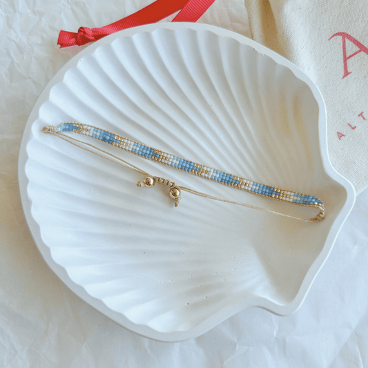 Pulsera Artesanal Tejida de Mostacillas Francisca en tonos azul y dorado, con cierre ajustable en hilo beige, sobre una concha blanca. Diseño hecho a mano con mostacillas de alta calidad.