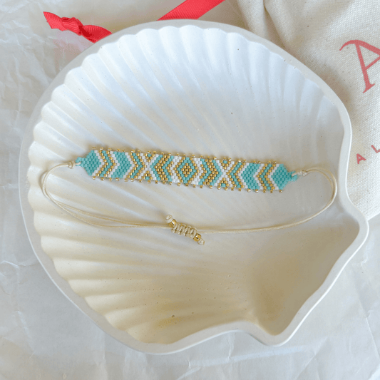Pulsera Artesanal Tejida de Mostacillas Inés en tonos turquesa, blanco y dorado, con patrón geométrico y cierre ajustable en hilo beige, sobre una concha blanca.