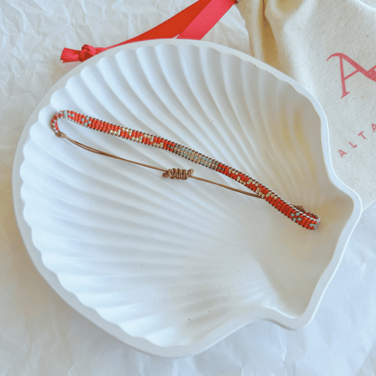 Pulsera Artesanal Tejida de Mostacillas Javiera en tonos rojo, naranja y dorado, con cierre ajustable en hilo marrón, sobre una concha blanca. Diseño hecho a mano con mostacillas de alta calidad.