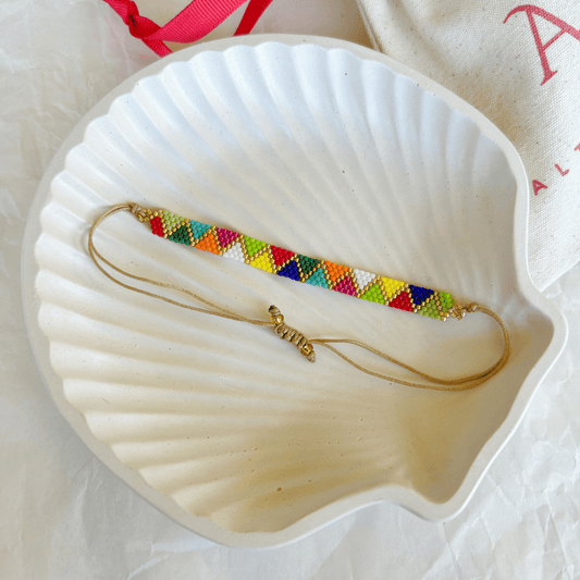 Pulsera Artesanal Tejida de Mostacillas Laura con diseño geométrico multicolor, cierre ajustable en hilo beige y detalles dorados, sobre una concha blanca.