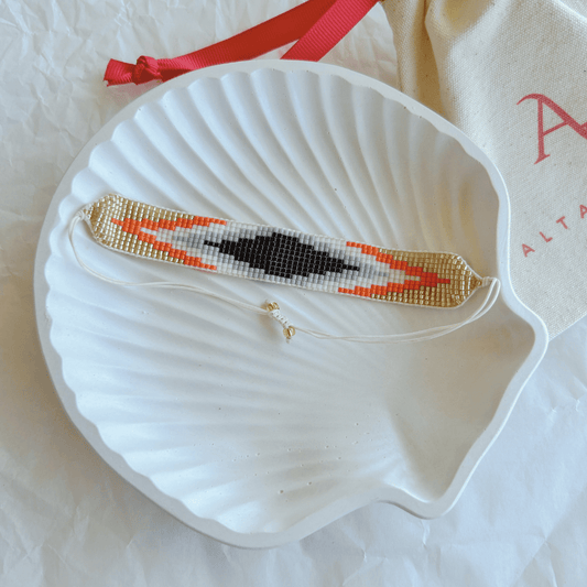 Pulsera Artesanal Tejida de Mostacillas Loreto en tonos negro, blanco, naranja y dorado, con cierre ajustable en hilo blanco, sobre una concha blanca. Accesorio elegante y sofisticado.