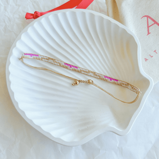 Pulsera Artesanal Tejida de Mostacillas Lucía en tonos rosa, dorado y verde menta, con cierre ajustable en hilo beige y detalles dorados, sobre una concha blanca.