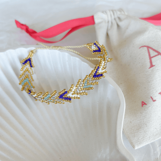 Pulsera Artesanal Tejida de Mostacillas María Gracia en tonos azul y blanco, con detalles dorados y diseño sofisticado, sobre una concha blanca.