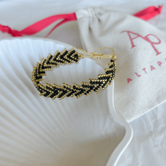 Pulsera Artesanal Tejida de Mostacillas María Gracia en tonos negro y dorado, con patrón trenzado y cierre ajustable, sobre una concha blanca.