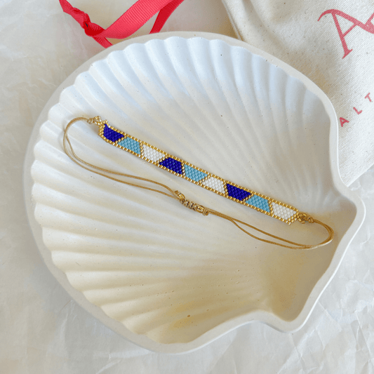 Pulsera Artesanal Tejida de Mostacillas Milagros en tonos azul marino, celeste, blanco y dorado, con patrón geométrico y cierre ajustable en hilo beige, sobre una concha blanca.