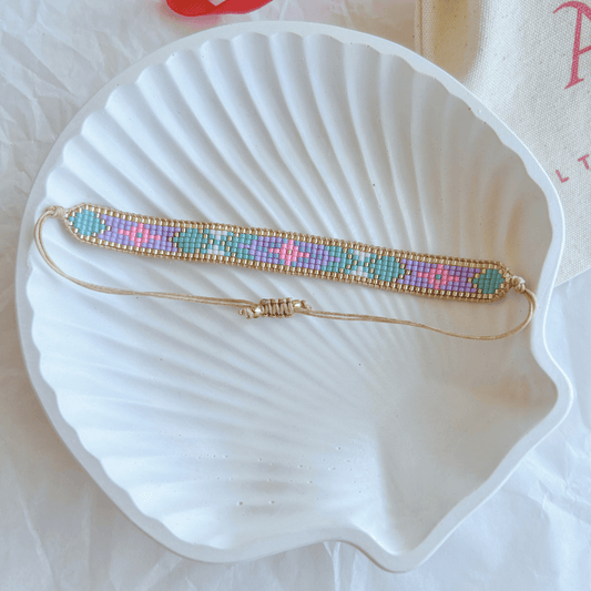 Pulsera Artesanal Tejida de Mostacillas Olivia en tonos turquesa, lila, rosa y dorado, con patrón geométrico y cierre ajustable en hilo beige, sobre una concha blanca.