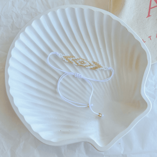 Pulsera Artesanal Tejida de Mostacillas Pola en tonos blanco y dorado, con diseño de ojo y cierre ajustable en hilo blanco, sobre una concha blanca.