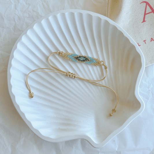 Pulsera Artesanal Tejida de Mostacillas Pola en tonos turquesa y negro, con diseño de ojo y cierre ajustable en hilo beige, sobre una concha blanca.