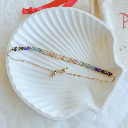 Pulsera Artesanal Tejida de Mostacillas Sofía en tonos degradados multicolor con detalles dorados, con cierre ajustable en hilo beige, sobre una concha blanca.