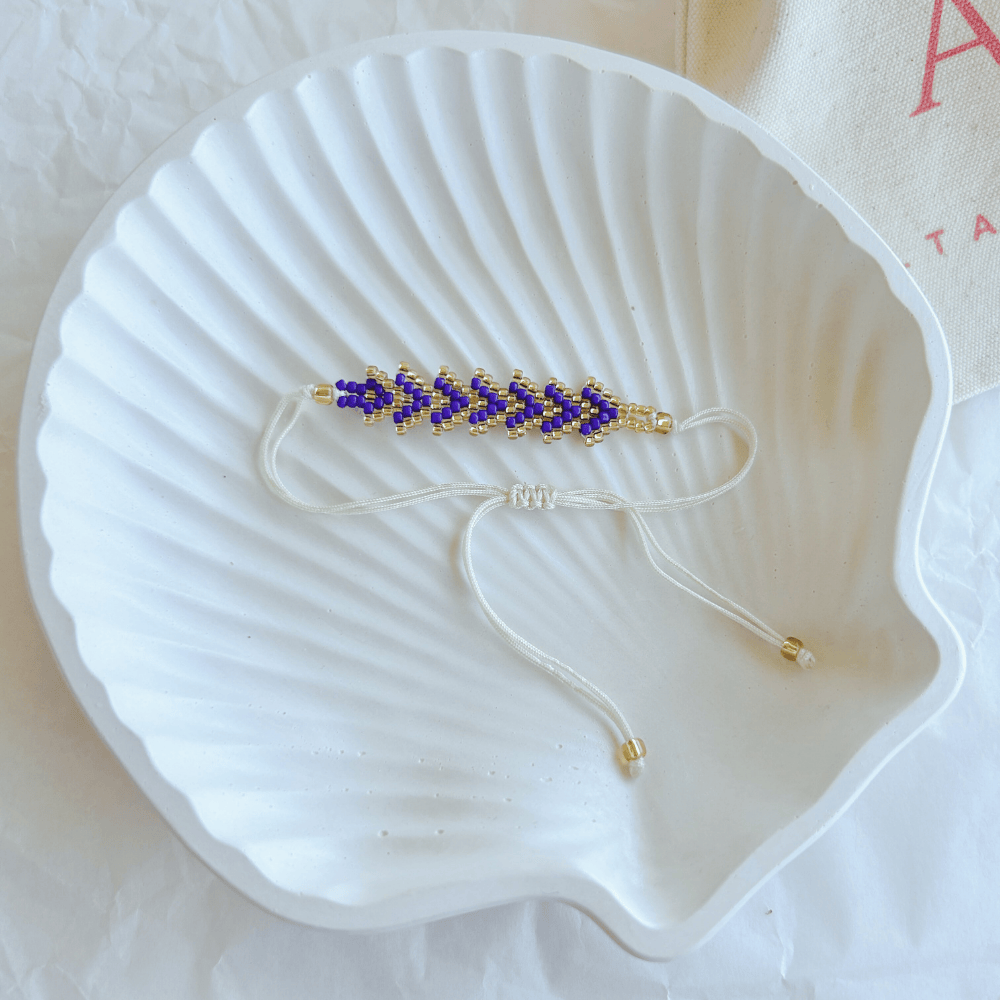 Pulsera Artesanal Tejida de Mostacillas Tere en azul rey con dorado, con cierre ajustable en hilo beige, sobre una concha blanca.
