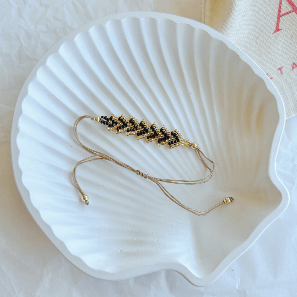 Pulsera Artesanal Tejida de Mostacillas Tere en negro con dorado, con cierre ajustable en hilo beige, sobre una concha blanca.