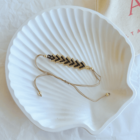 Pulsera Artesanal Tejida de Mostacillas Tere en negro con dorado, con cierre ajustable en hilo beige, sobre una concha blanca.
