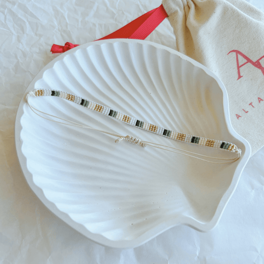 Pulsera Artesanal Tejida de Mostacillas Victoria en tonos blancos, verdes y dorados, con cierre ajustable en hilo beige, sobre una concha blanca.