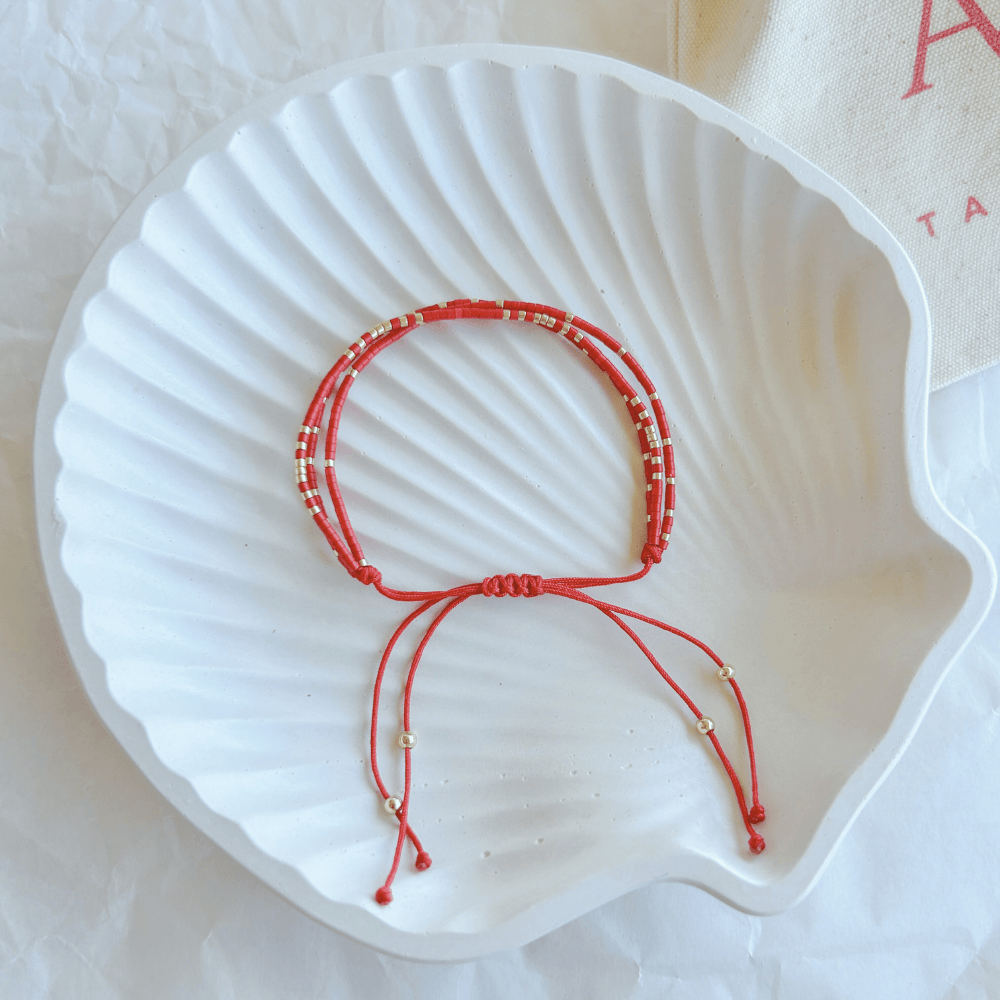 Pulsera Artesanal Triple Isidora sobre una concha blanca, mostrando su diseño artesanal de tres hileras en tonos rojo, dorado y turquesa, con cierre ajustable.