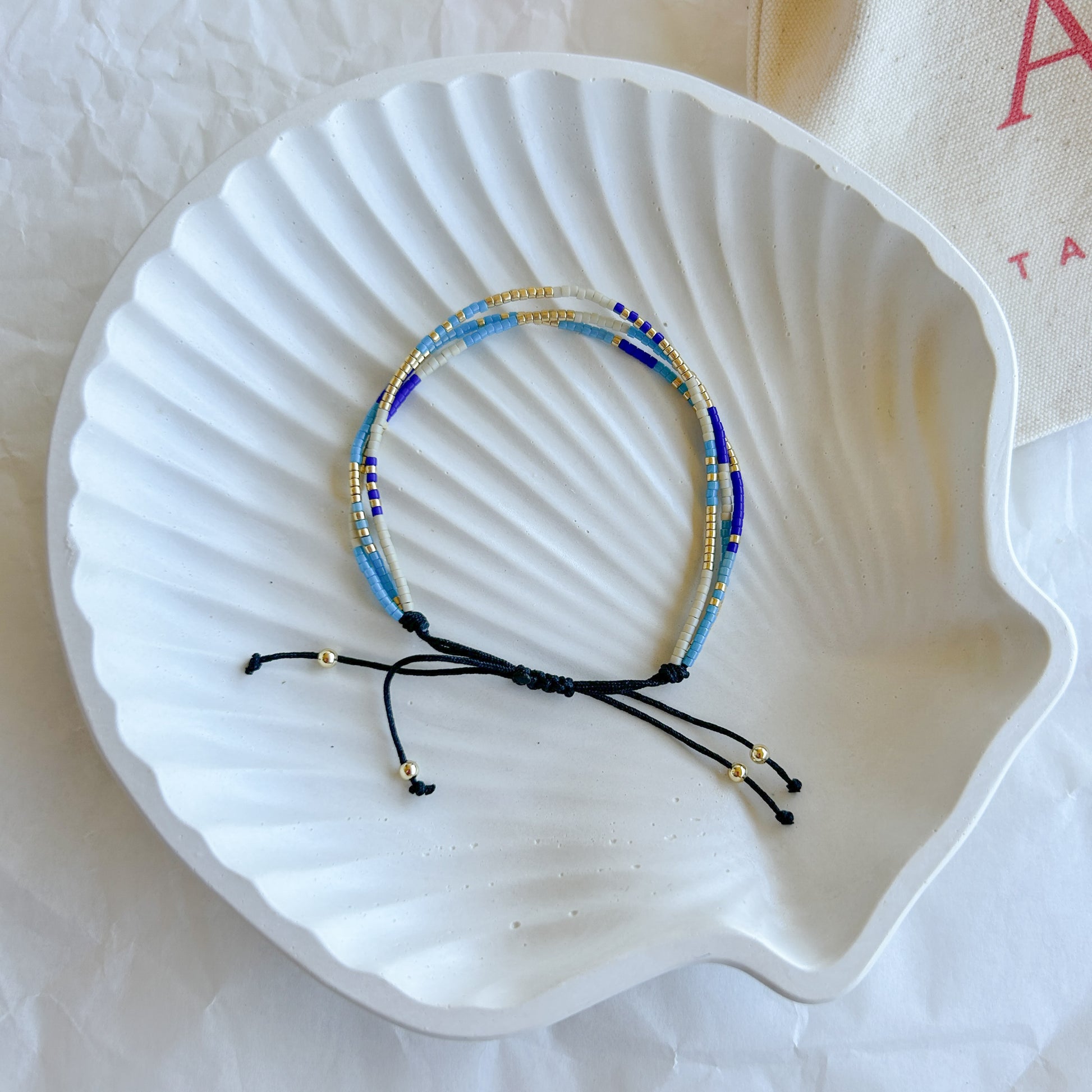 Pulsera Artesanal Triple Martina sobre una concha blanca, destacando su diseño de tres hileras en tonos azul, dorado y blanco con cierre ajustable.
