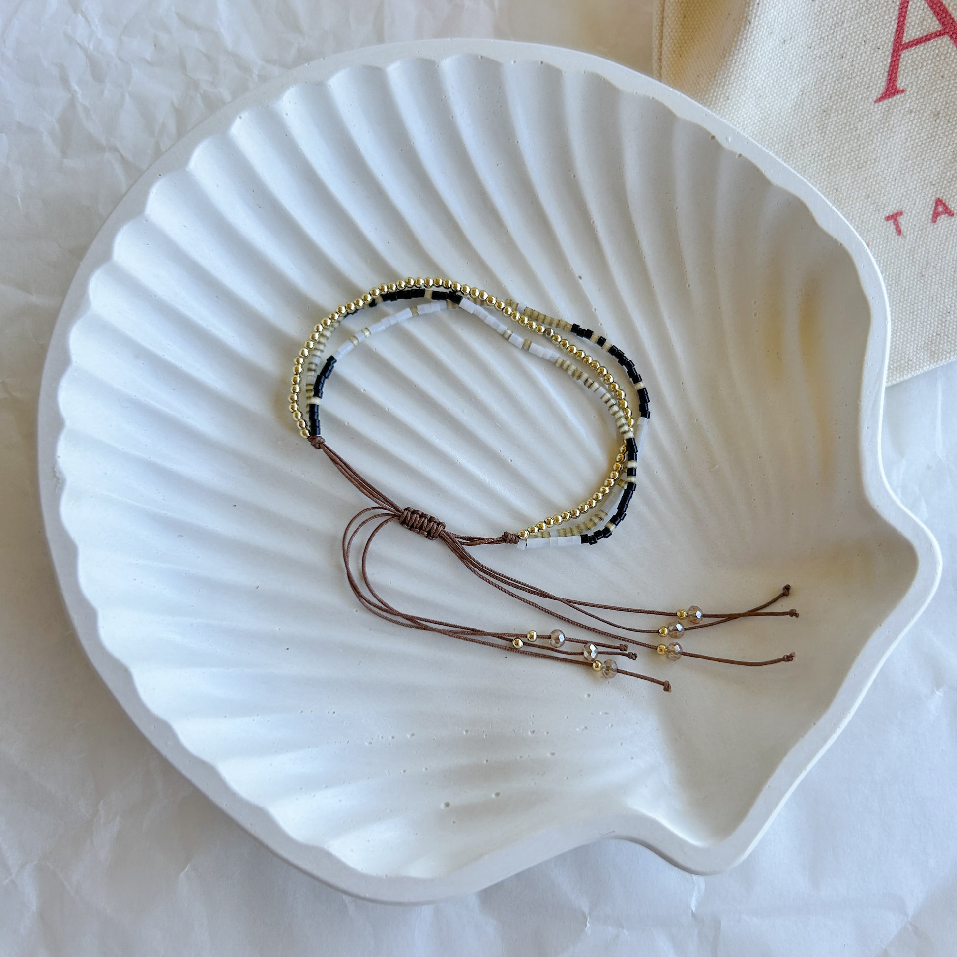 Pulsera Artesanal Triple Matilde sobre una concha blanca, resaltando su diseño de tres hileras en negro, dorado y blanco con cierre ajustable.