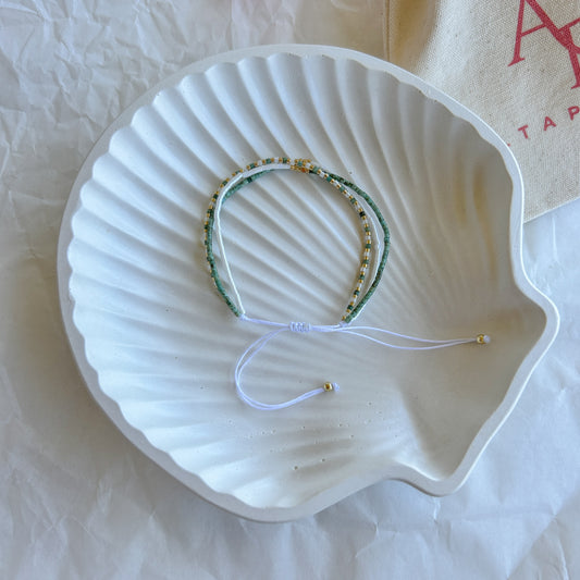 Pulsera Artesanal Triple Rafaela sobre una concha blanca, con un diseño artesanal de tres hileras en verde, blanco y dorado con cierre ajustable.