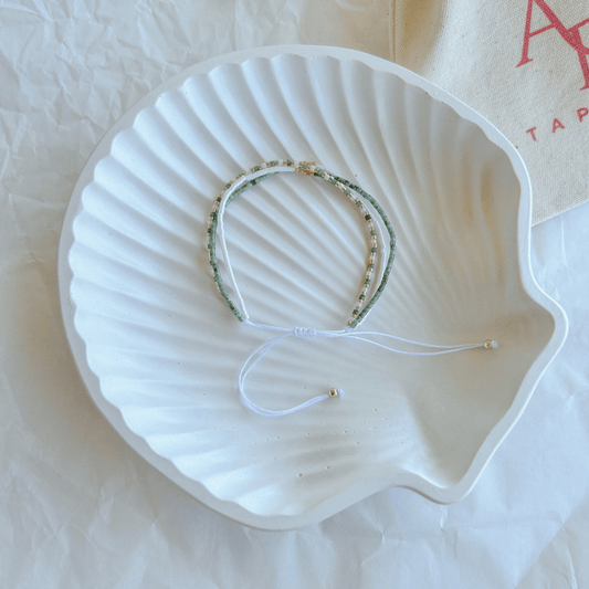 Pulsera Artesanal Triple Rafaela sobre una concha blanca, con un diseño artesanal de tres hileras en verde, blanco y dorado con cierre ajustable.