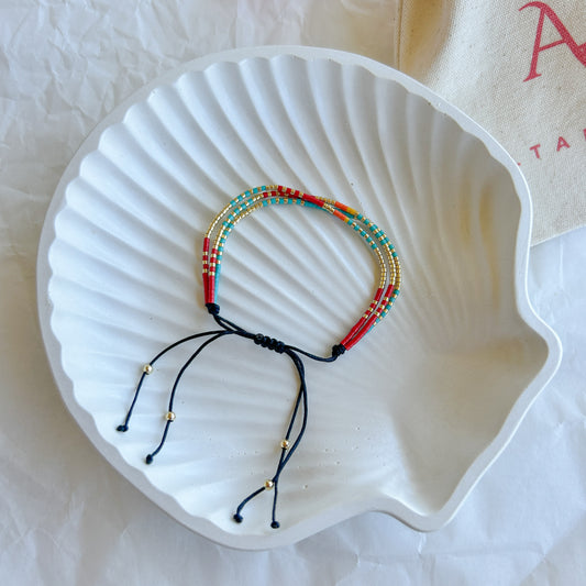 Pulsera Artesanal Triple Trinidad sobre una concha blanca, mostrando su diseño artesanal de tres hileras en tonos rojo, turquesa y dorado con cierre ajustable.
