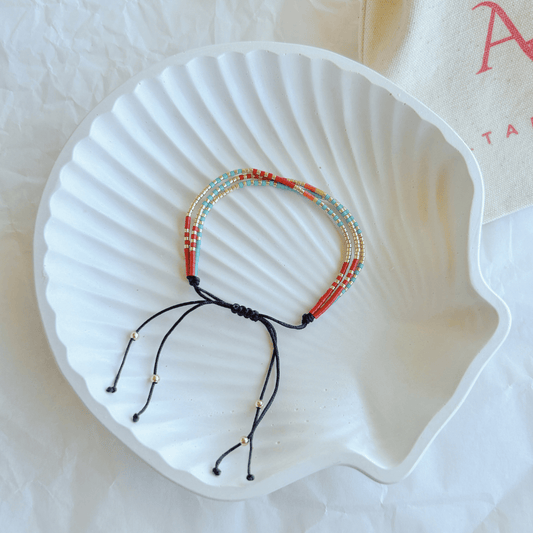 Pulsera Artesanal Triple Trinidad sobre una concha blanca, mostrando su diseño artesanal de tres hileras en tonos rojo, turquesa y dorado con cierre ajustable.